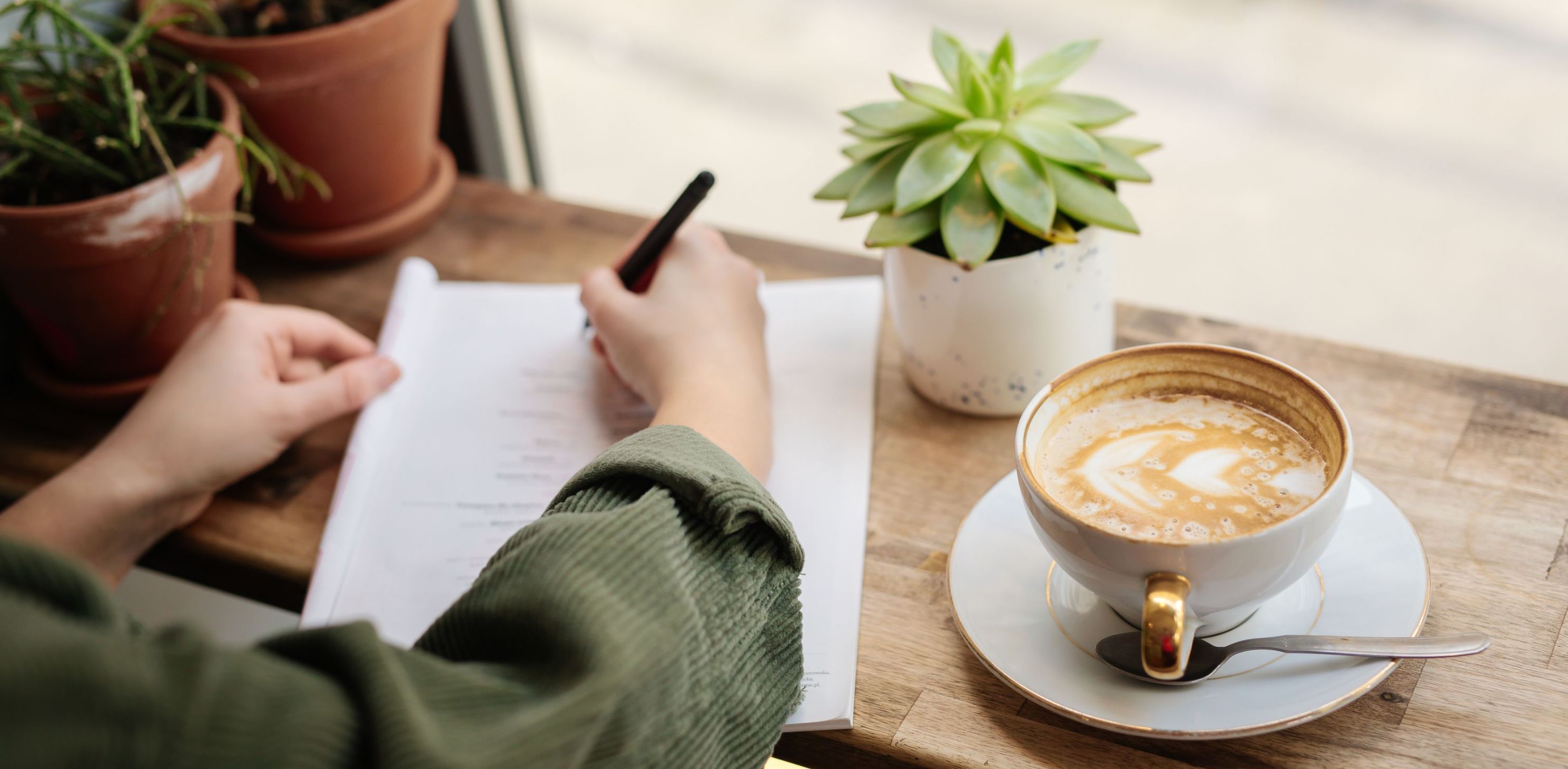 Person skriver på papirer med kaffekopp og planter på bordet